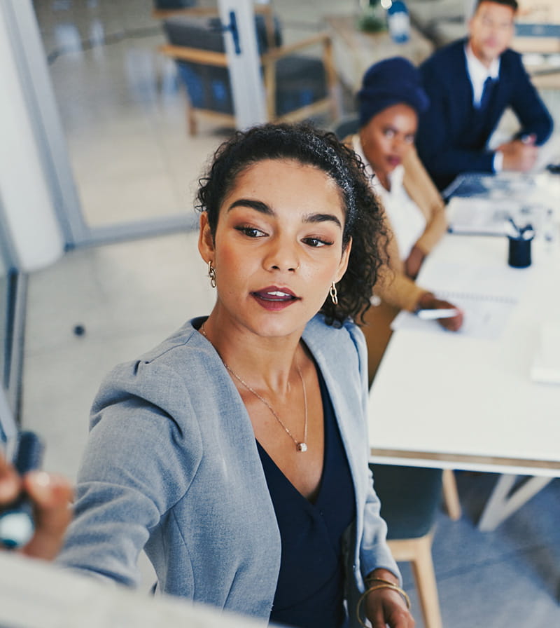 Female associate writing on a whiteboard with other employees sitting at a table in conference